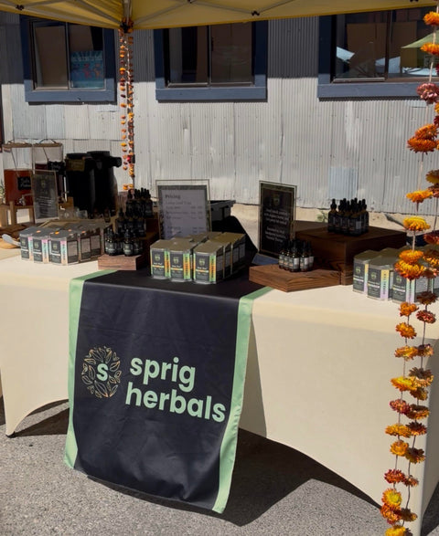 A market booth for 'sprig herbals' under a yellow canopy, featuring a cream tablecloth, a black table runner with the brand logo, and various packaged herbal products displayed on tables.