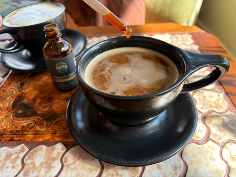 Drops of Sunnyside Spice Herbal Extract being added to a cup of hot coffee on a tiled table.