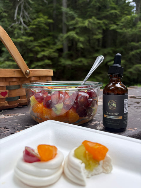 Bottle of Sprig Herbals Chamomile Extract beside fruit salad and small pastries on a picnic table.